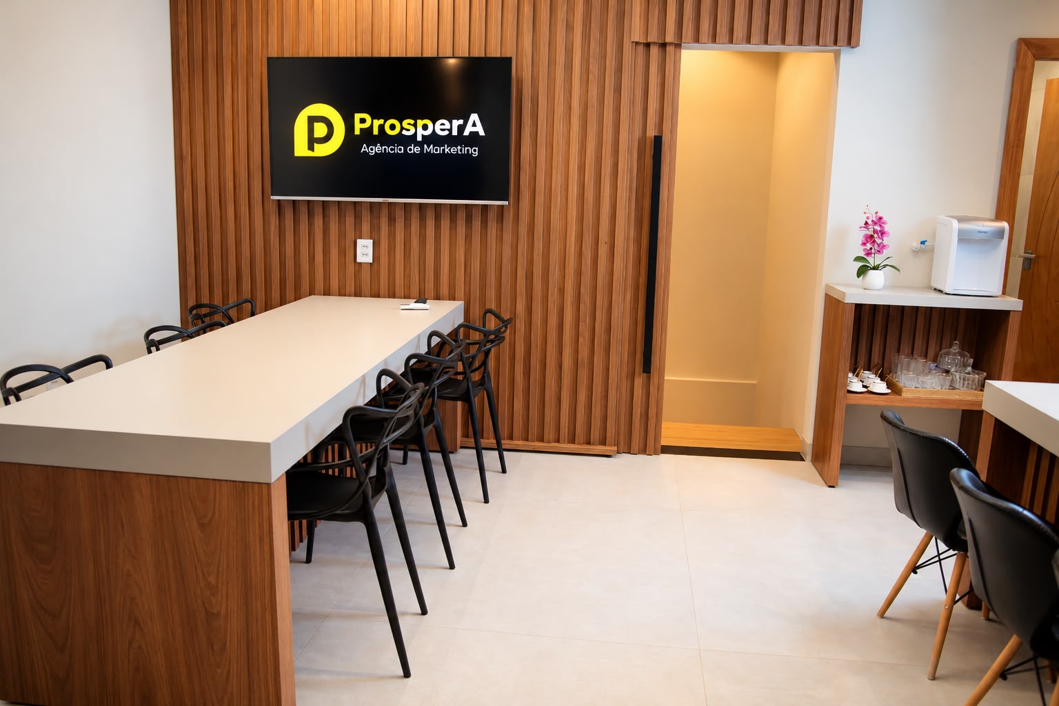 Área de convivência com mesa de madeira e TV exibindo a marca Prospera